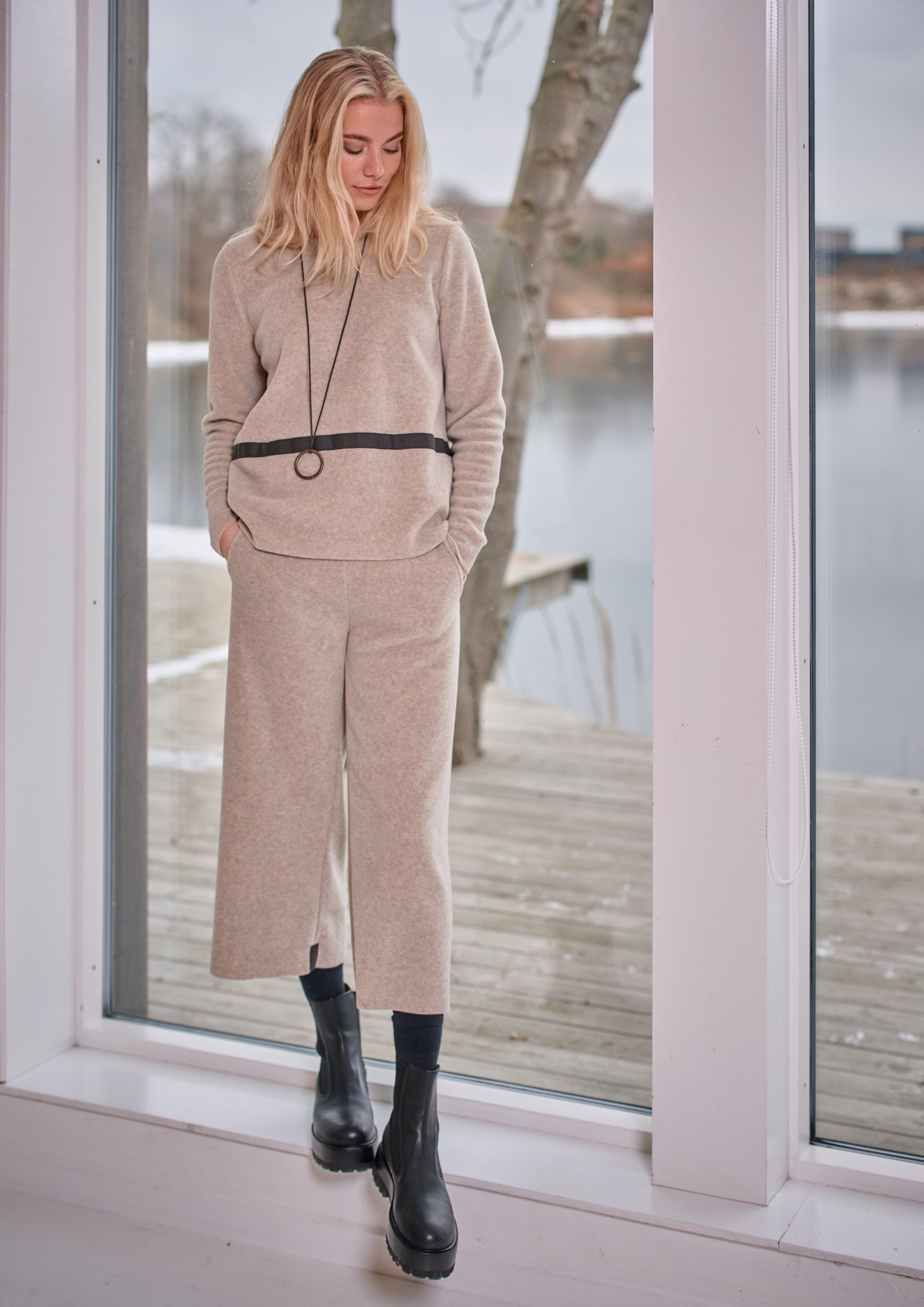 Woman in beige tracksuit standing by a window with a view of water and trees.