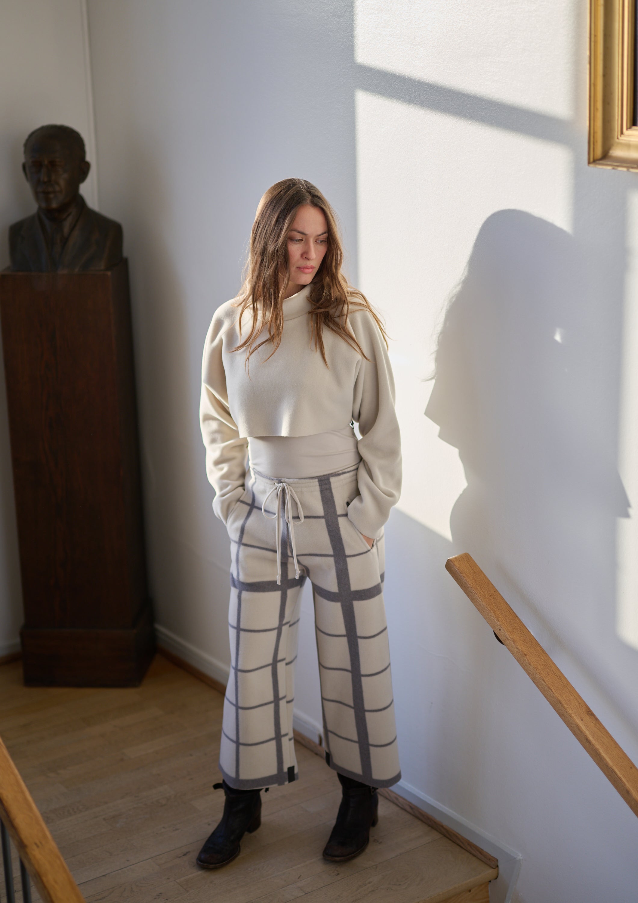 Woman in a beige sweater and checkered pants standing in a room with a sculpture and window.