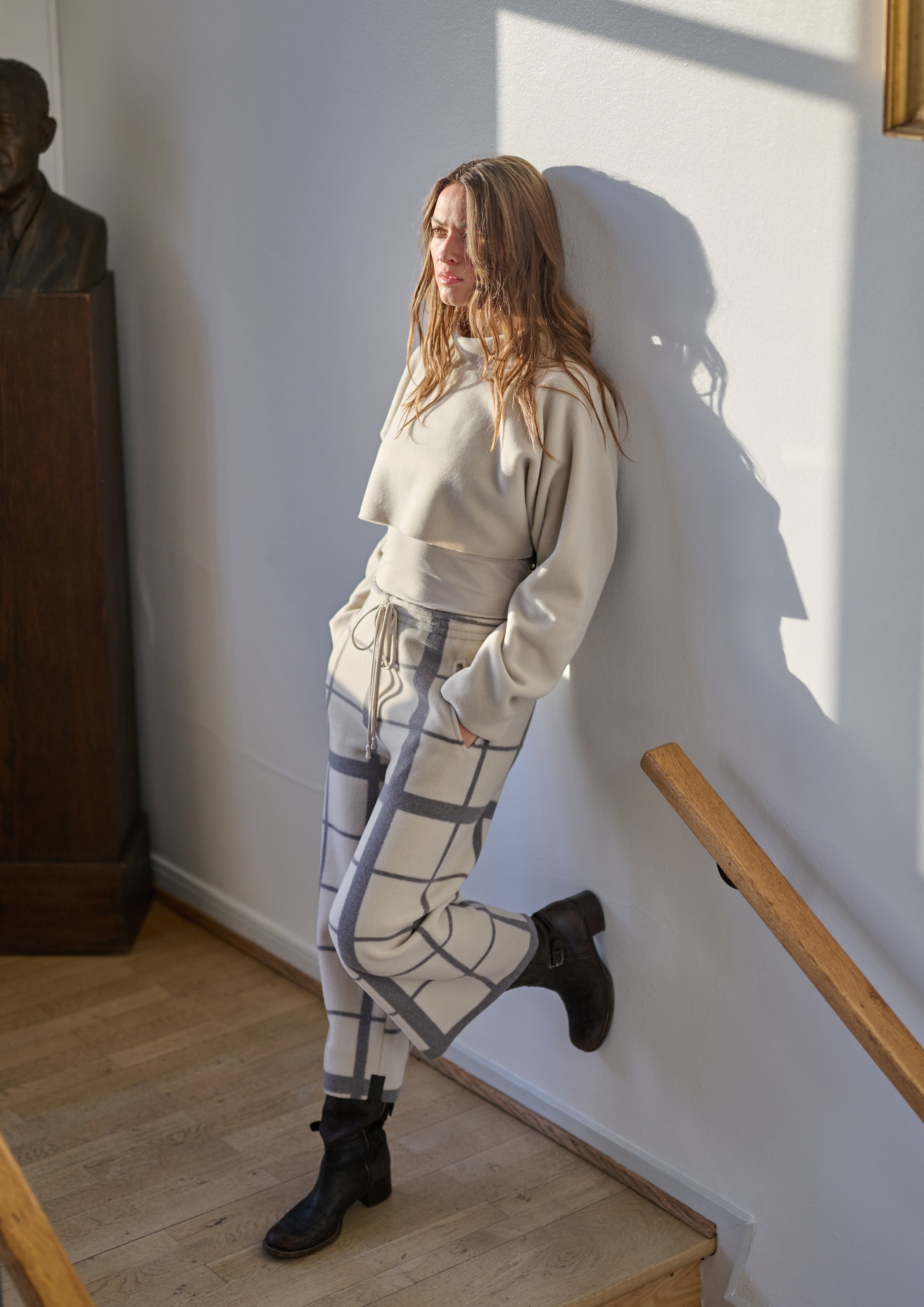 Woman in a light gray outfit standing against a white wall with a wooden railing.