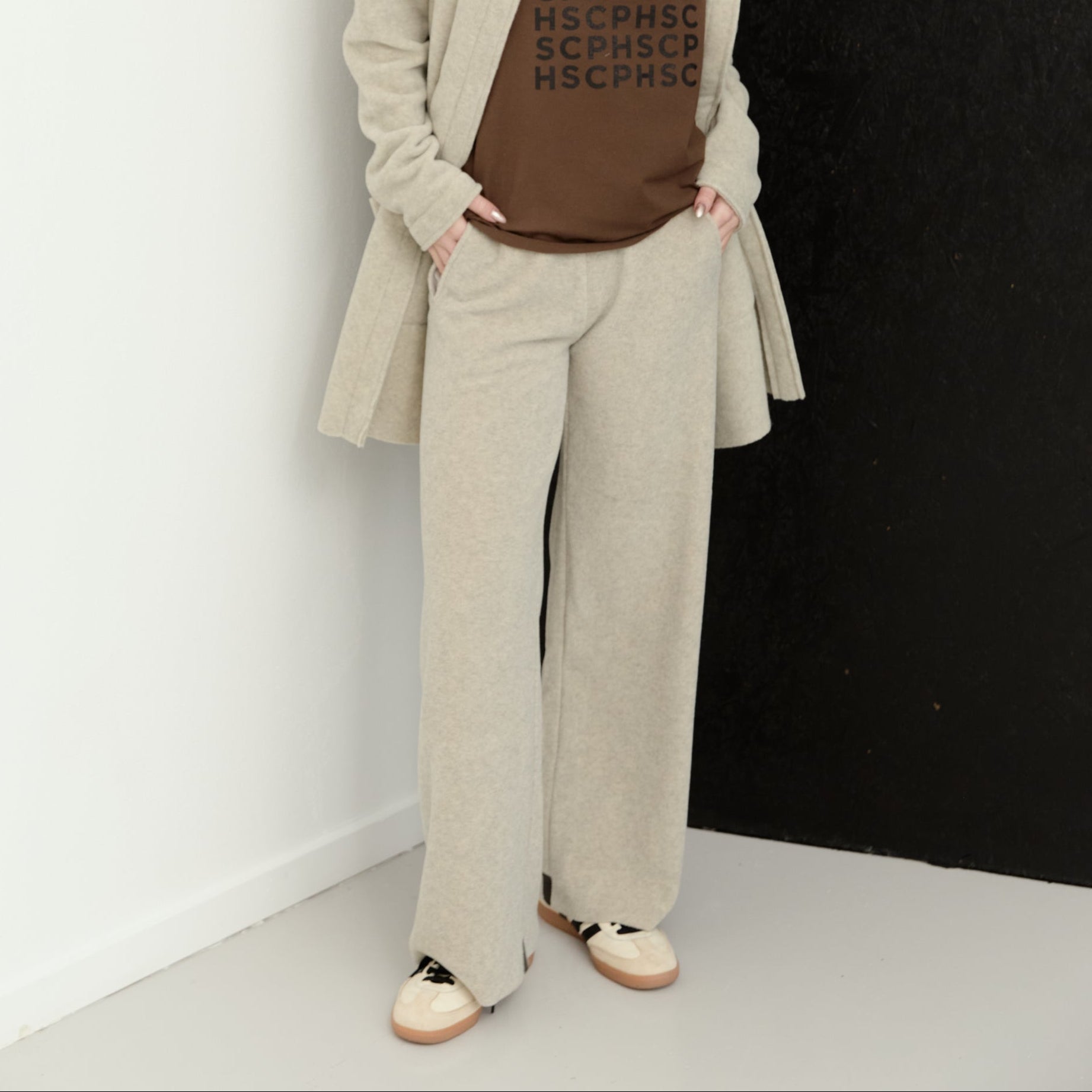A woman stands against a split white and black wall wearing a beige hooded cardigan, matching beige wide-leg trousers, a brown graphic T-shirt, and beige-and-white sneakers.