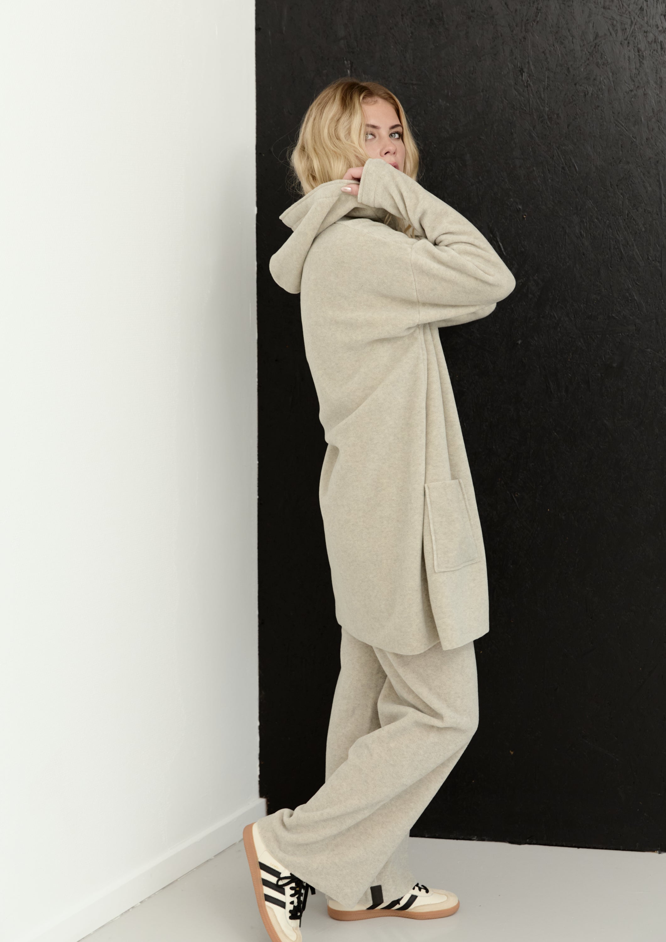 A woman wearing a matching beige hooded sweatshirt and wide-leg sweatpants stands in a studio corner with a white wall on the left and a black wall on the right.