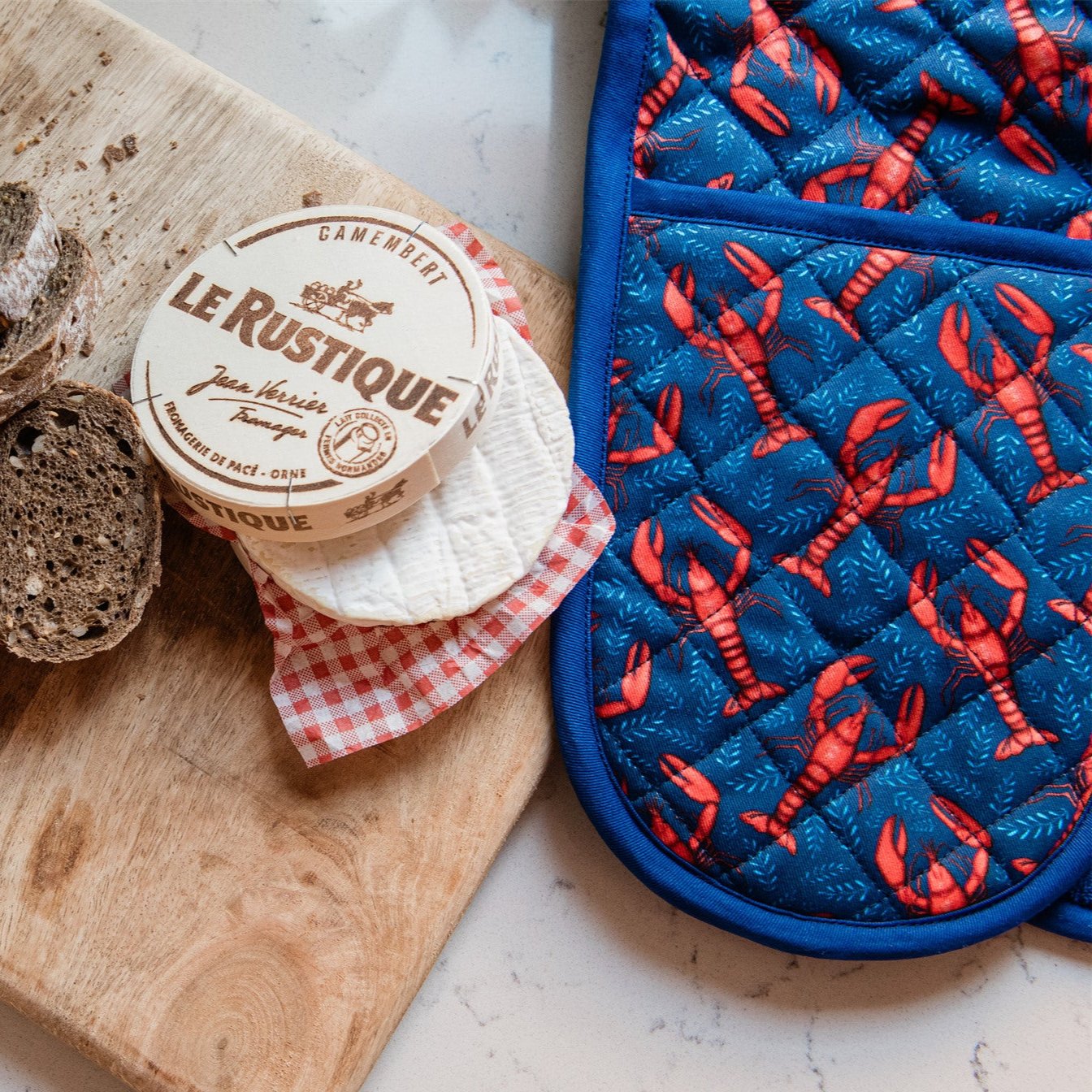 Le Rustique Camembert cheese rounds on a wooden board, next to a blue quilted oven mitt with a lobster print.