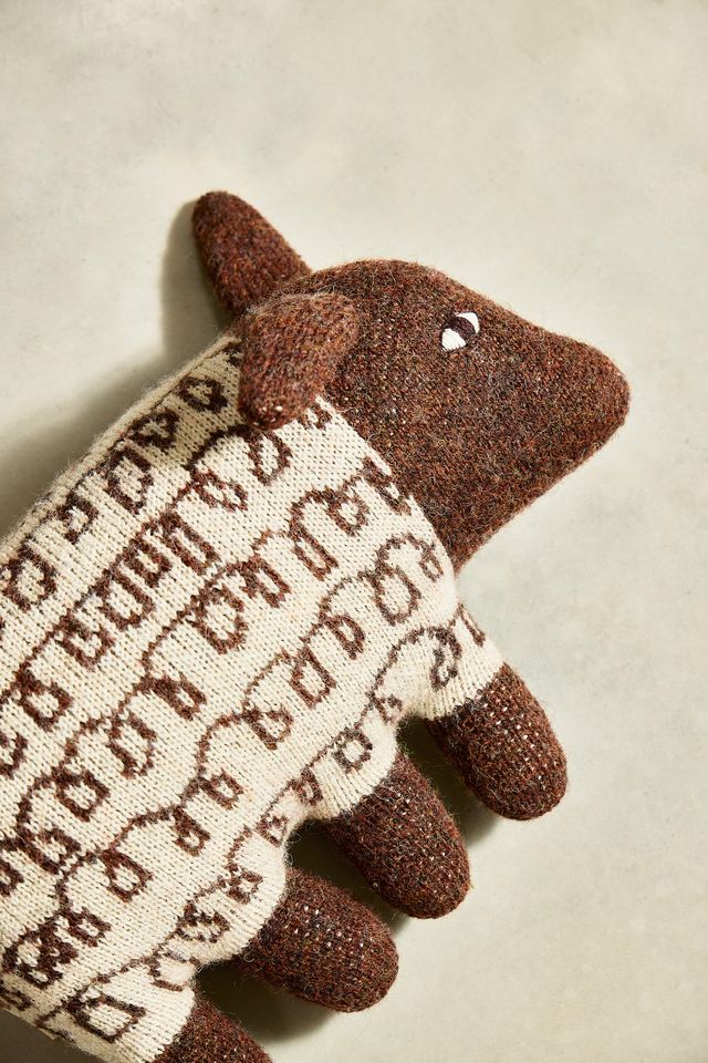A close-up of a brown knit sheep-shaped accessory, likely a glove or pouch, featuring a cream knit body with brown lettering and a small white eye detail, set against a neutral beige background.