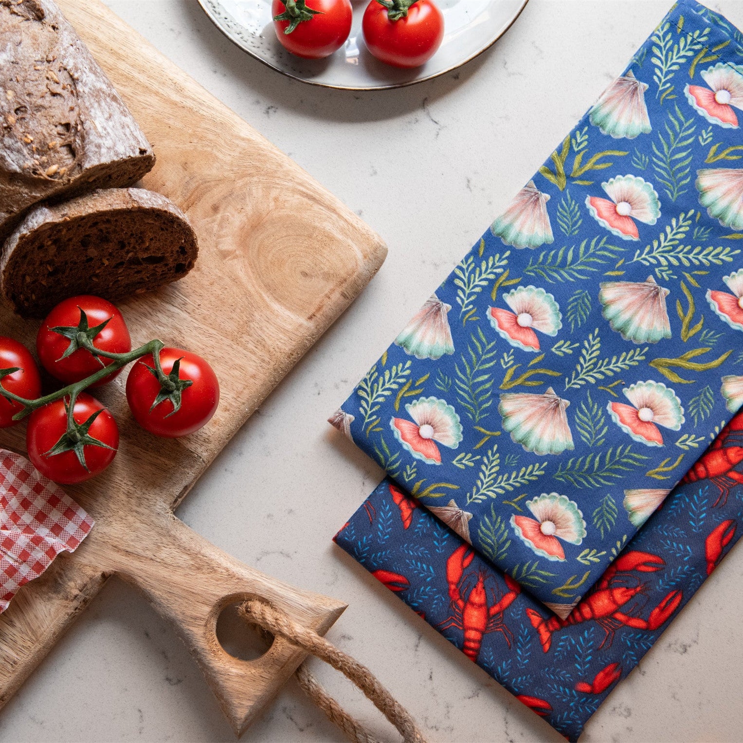 Two kitchen tea towels; one features a seashell and foliage pattern on a dark blue background, the other shows red lobsters