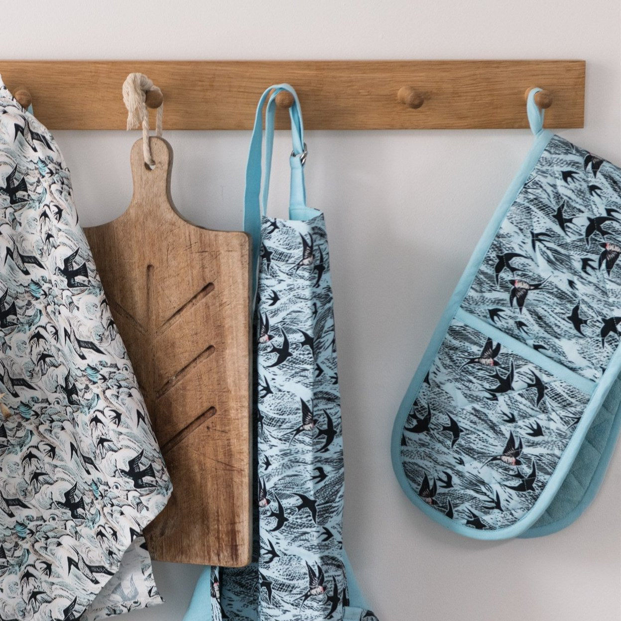 Wooden cutting board and light blue kitchen textiles with a bird print hanging on a wall-mounted rack.