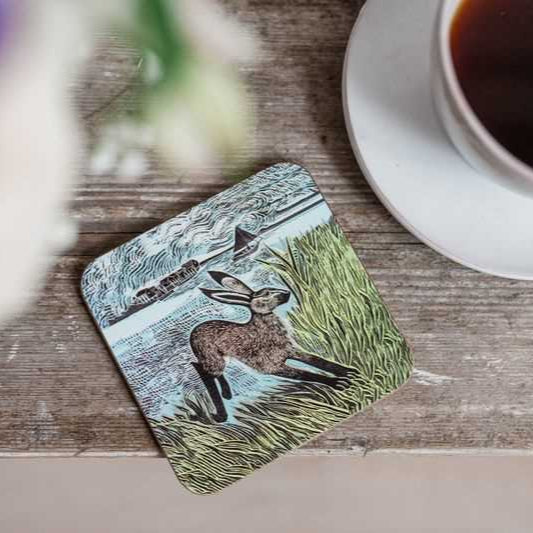 Square coaster featuring a linocut-style illustration of a hare in tall grass, with a distant train and sailboat visible in