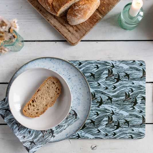 Blue placemat with a repeating pattern of dark birds in flight, shown with a light blue speckled dinner plate and bowl