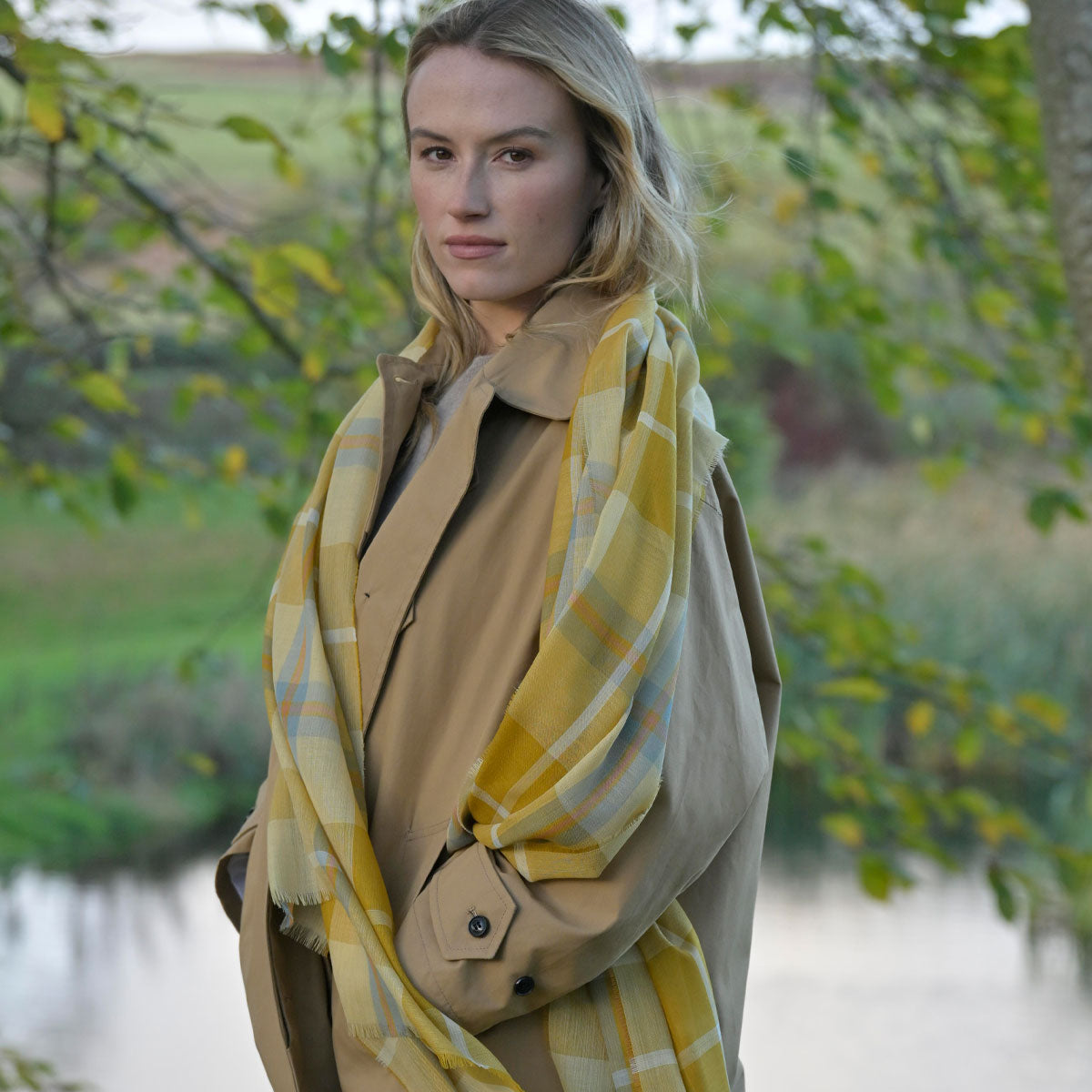 Woman wearing a yellow plaid scarf and beige coat outdoors with greenery in the background