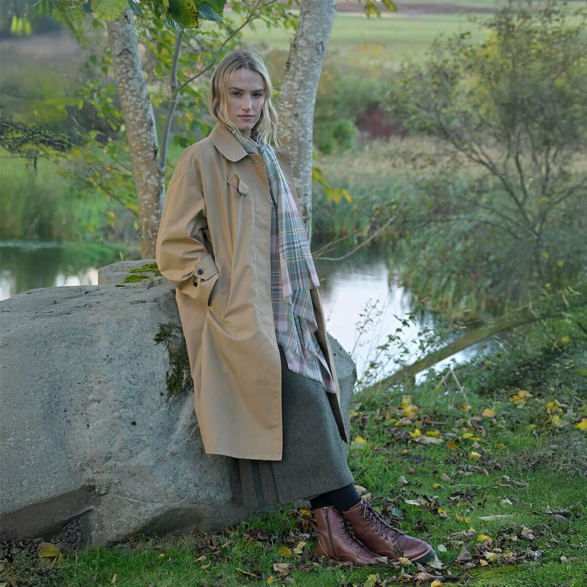 Woman in a beige trench coat standing by a pond with trees and grass in the background