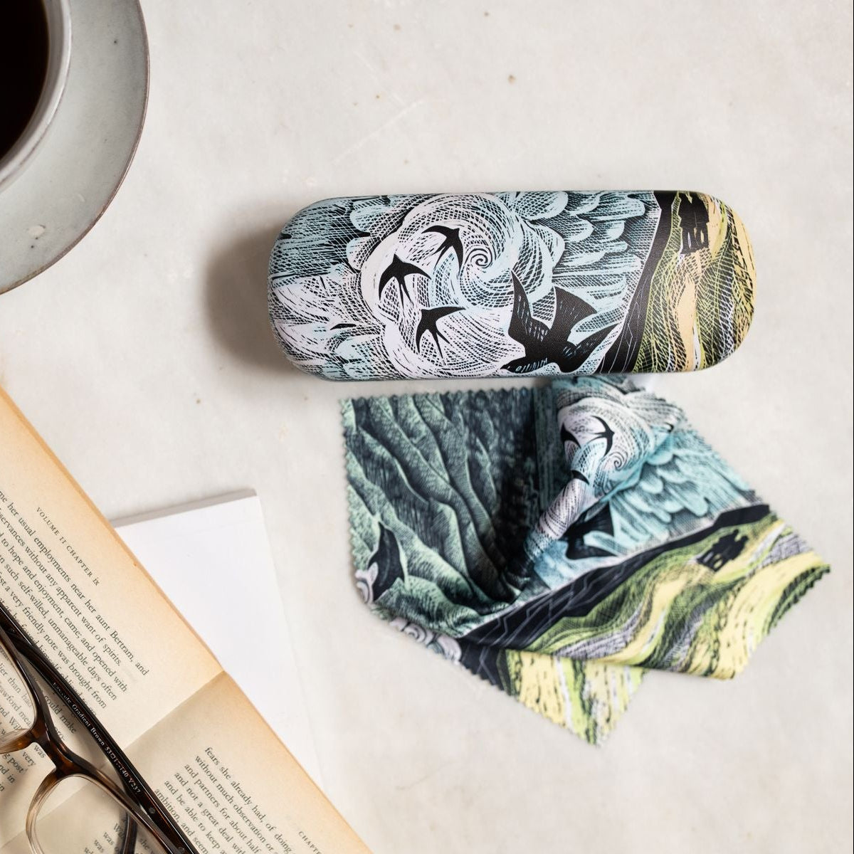 A pair of eyeglasses with a blue, green, and white patterned fabric case and matching cloth, styled in a flat-lay with a cup of coffee, an open book, and a pair of eyeglasses on a light surface.