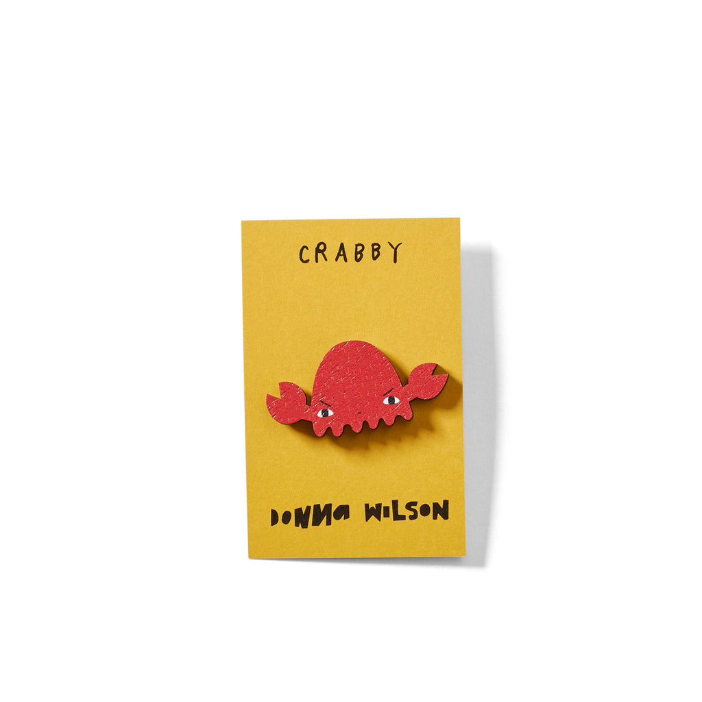 Red crab brooch on a yellow card featuring the text "CRABBY" and "DONNA WILSON" below.