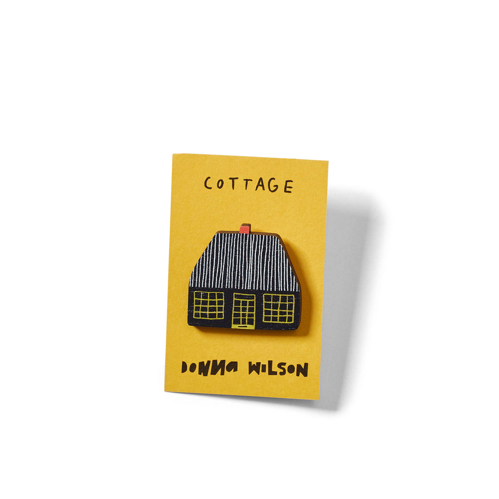 A cottage pin in black with yellow windows and a red roof on a yellow card; by Donna Wilson.
