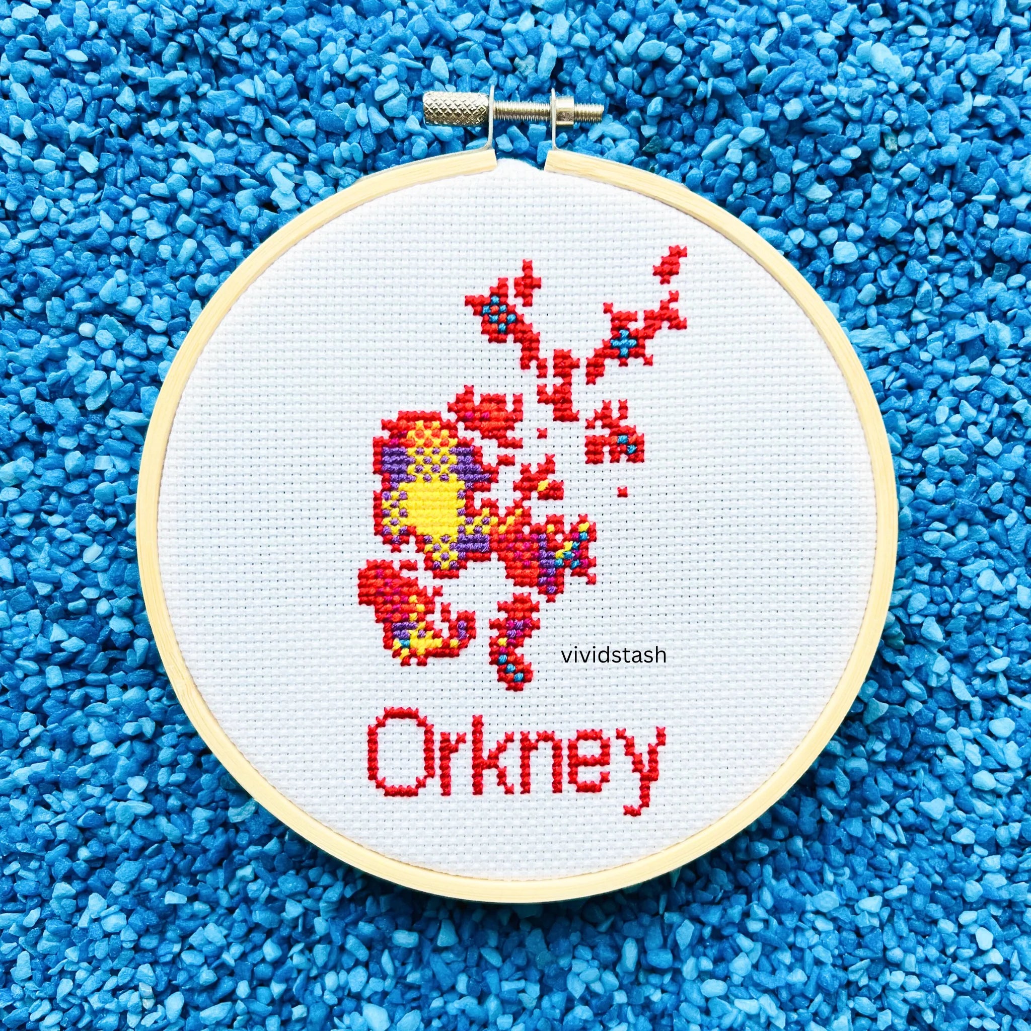 Cross-stitch embroidery in a wooden hoop depicting Orkney islands in red, yellow, and purple threads on white fabric.