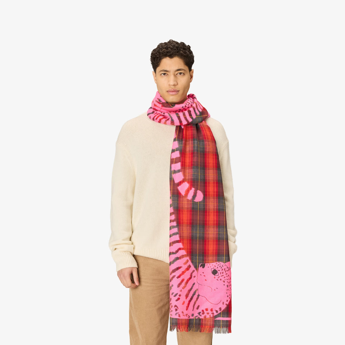 A model wearing a cream knit sweater, beige trousers, and a vibrant pink and red scarf featuring a tiger design and plaid pattern, set against a plain light gray background.