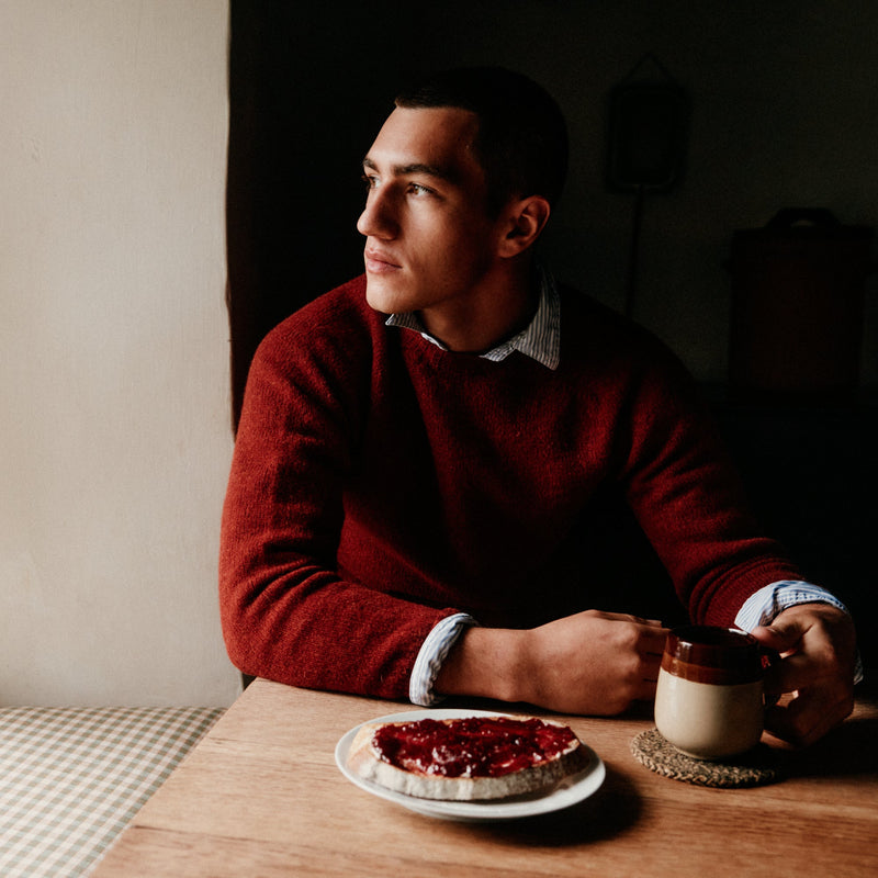 A man wearing a textured dark red knit sweater sits at a wooden table, looking off-camera. In front of him is a plate with a red fruit tart and a ceramic mug on a coaster.