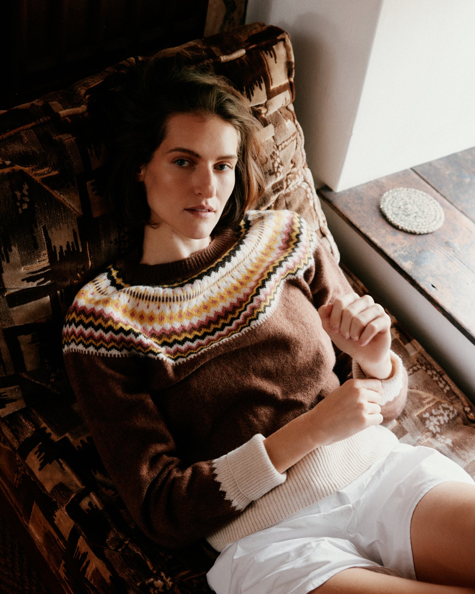 Woman wearing a patterned sweater sitting on a wooden chair by a window.