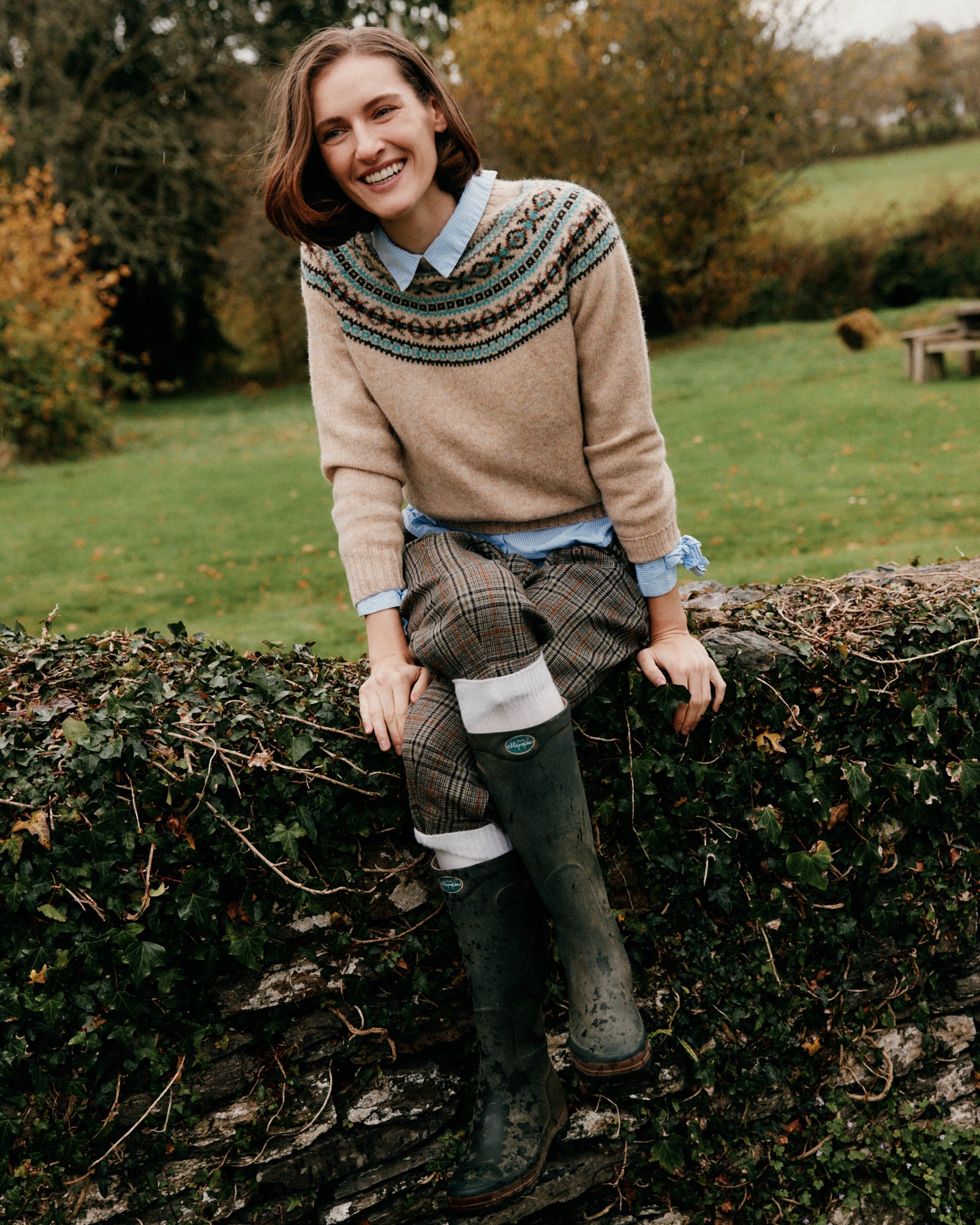 Woman in a patterned sweater and plaid skirt standing outdoors by a hedge.
