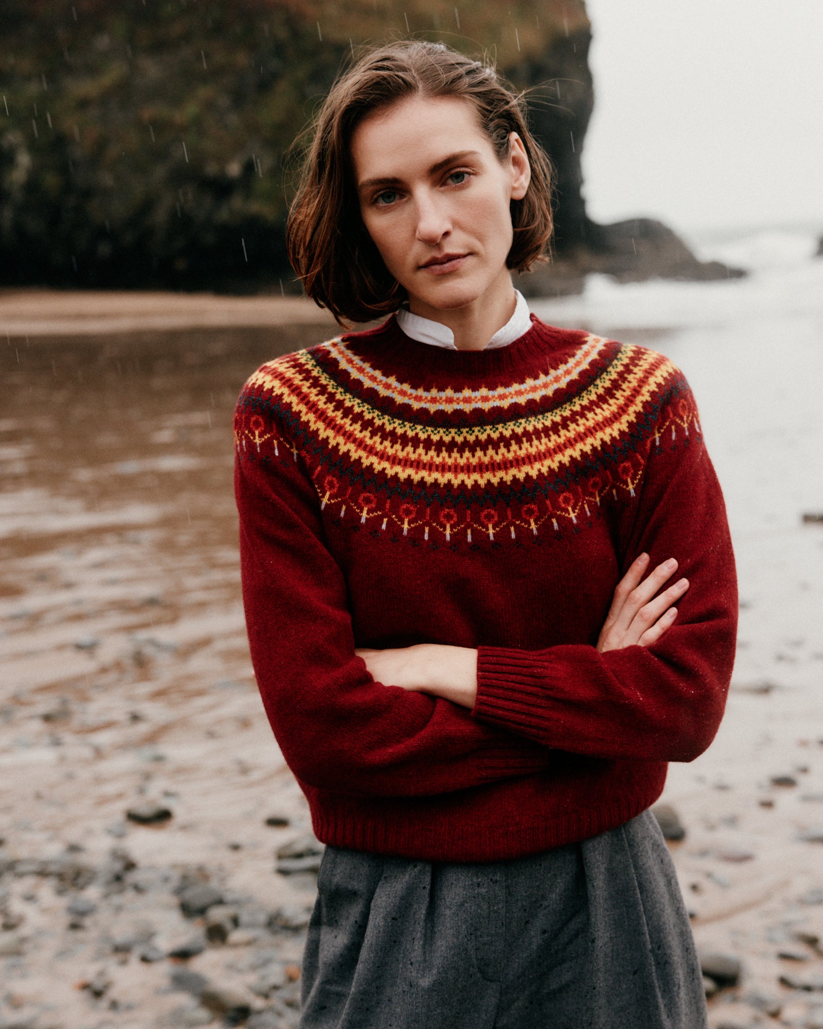 Woman wearing a red patterned sweater standing on a rocky shoreline.