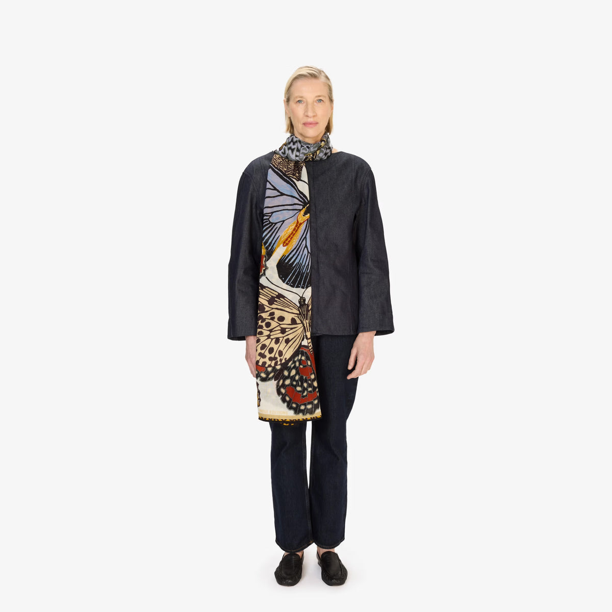 A model stands against a plain white studio background wearing a dark charcoal jacket, dark trousers, and a long scarf with a large butterfly print in cream, blue, orange, and red.