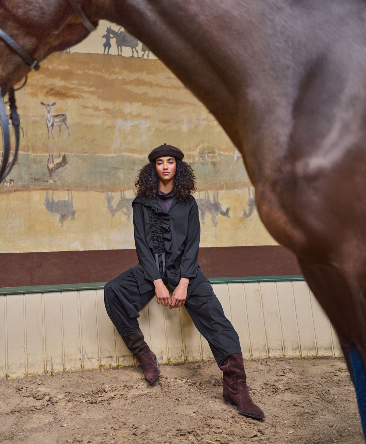A model wearing a black ruffled long-sleeve top, black wide-leg pants, and dark brown suede boots sits on a wooden barrier in an equestrian stable, framed by a large horse in the foreground.