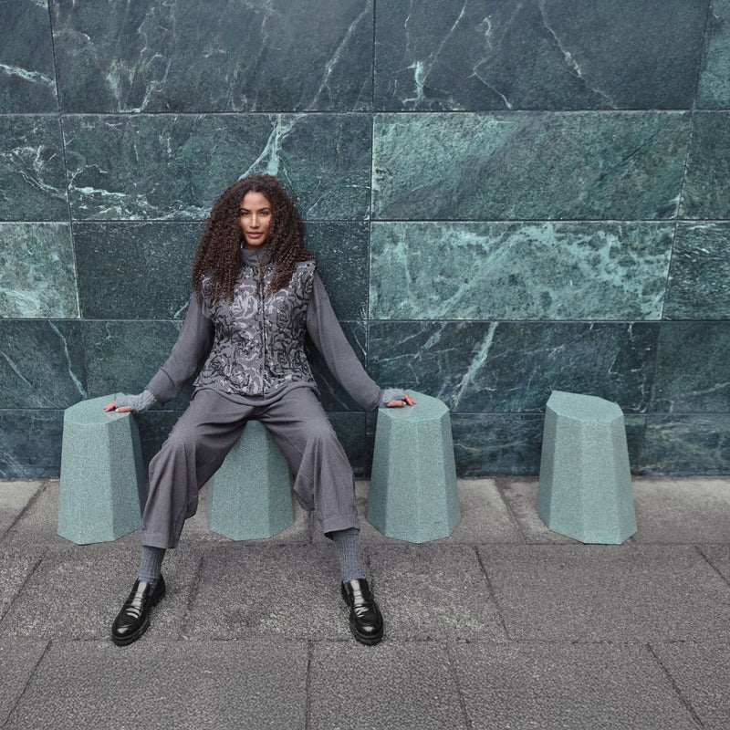 A woman sits on a light blue-green concrete bench in front of a dark green marble wall. She is wearing a gray patterned long-sleeve top and matching gray trousers with black shoes.