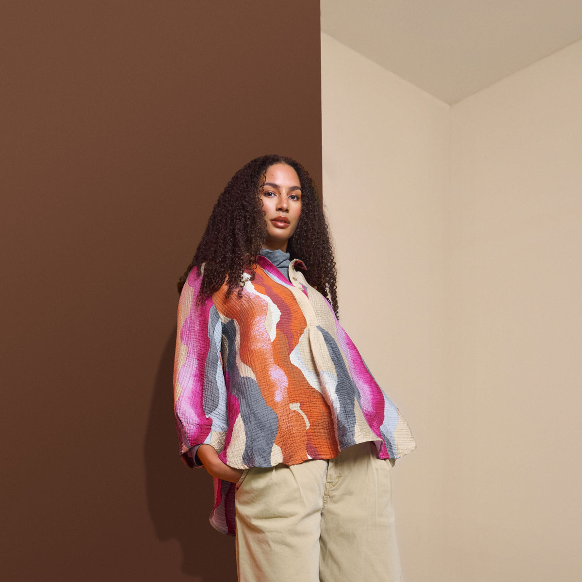 A model stands in a minimalist studio setting with a brown wall and cream-colored corner walls, wearing a colorful abstract print long-sleeve shirt, beige cropped pants, and dark brown knee-high boots.
