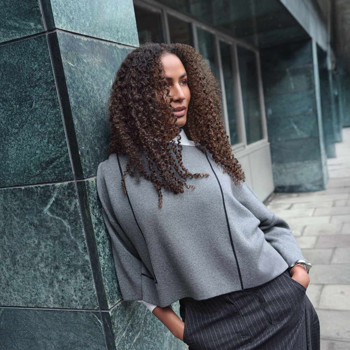 A woman with long, curly brown hair leans against a dark green marble column outdoors. She is wearing a light gray cropped sweater with black piping details and a dark gray pinstriped skirt.