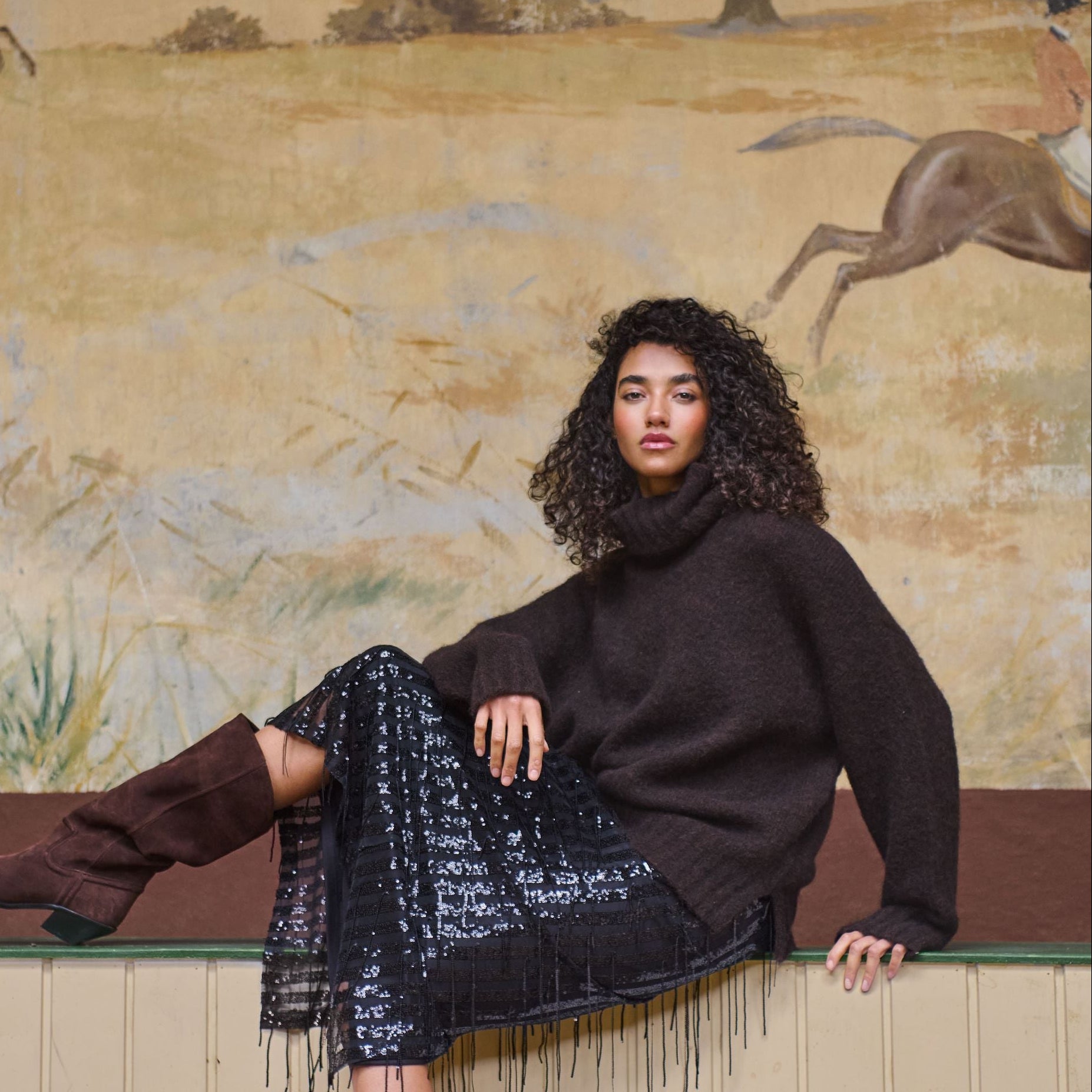 A woman with curly dark hair sits on a low wall in front of a painted equestrian scene, wearing a dark brown turtleneck sweater, a black fringed midi skirt, and dark brown knee-high boots.