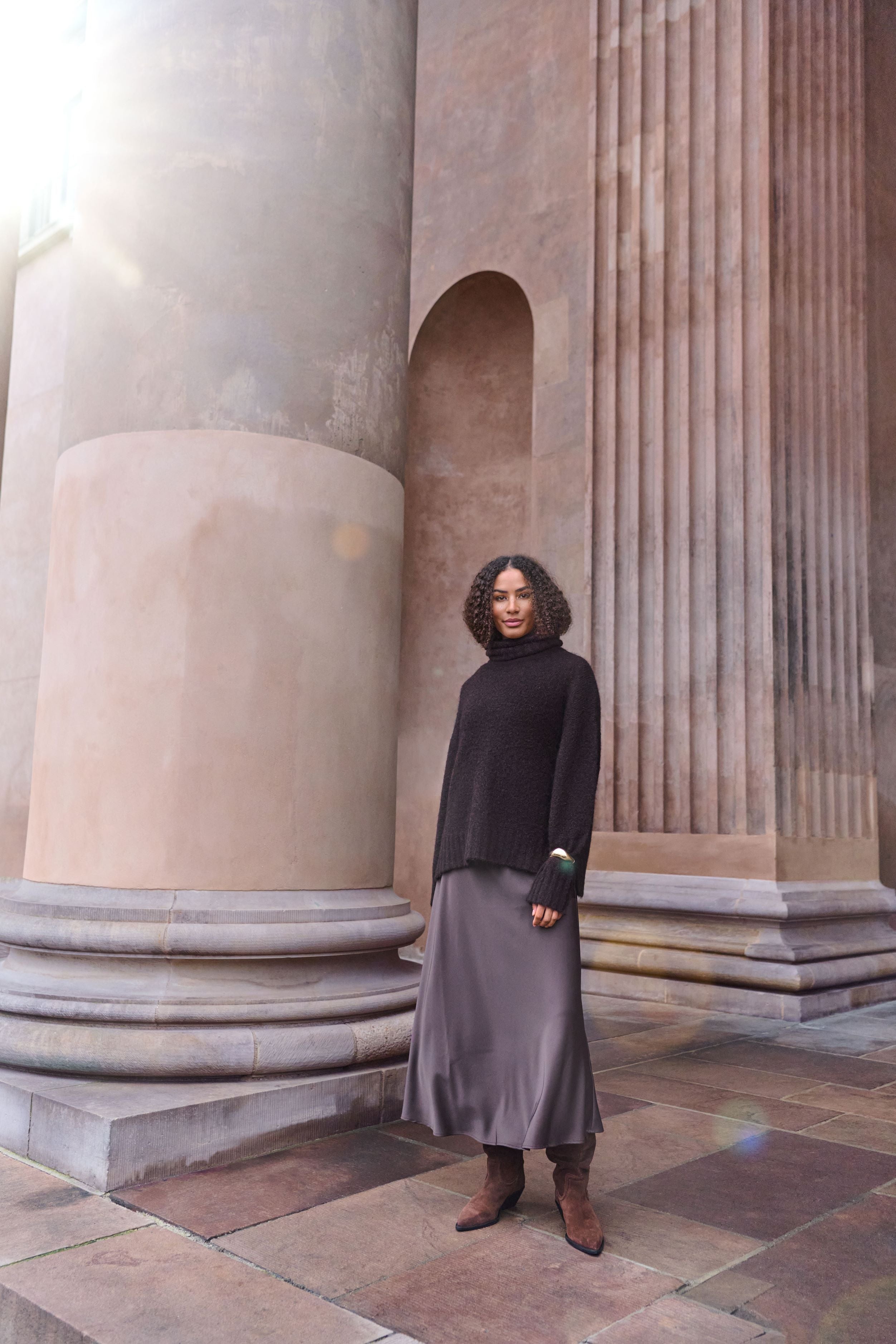 A woman stands in front of large classical stone columns, wearing a black turtleneck sweater, a long taupe-gray skirt, and brown ankle boots.