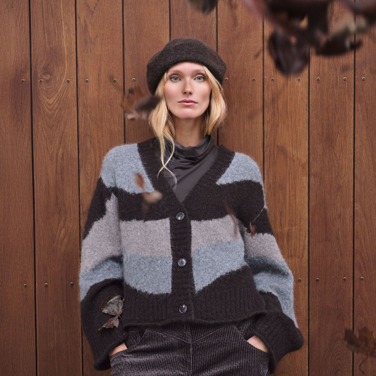 A woman wearing a dark gray and light gray striped knit cardigan, a dark beret, and a dark corduroy skirt, leaning against a wooden plank wall with autumn leaves in the foreground.