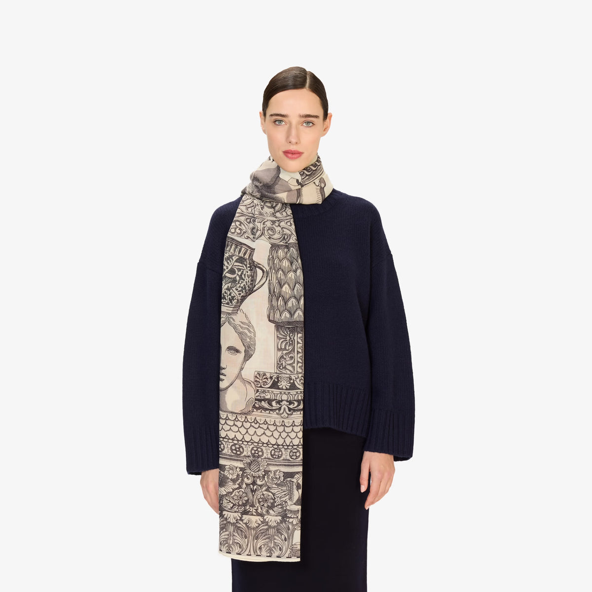 A model wearing a navy blue knit sweater and a long beige scarf with a black and gray classical bust and ornamental pattern, set against a plain light gray background.