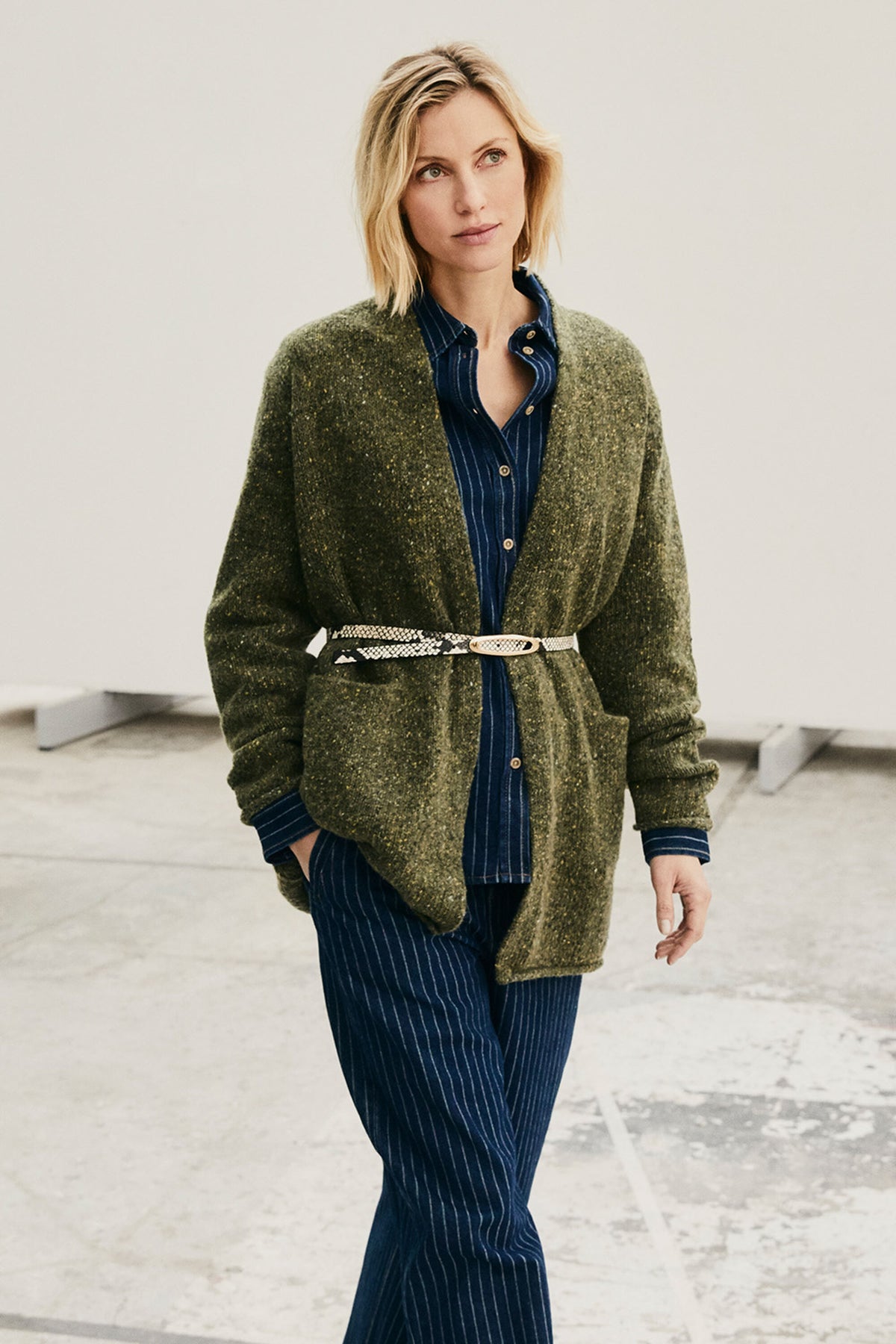 A woman modeling an olive green textured knit cardigan with front patch pockets, worn over a navy blue striped button-up shirt and paired with matching navy pinstriped trousers, a silver chain belt, and a gold buckle, in a minimalist studio setting.