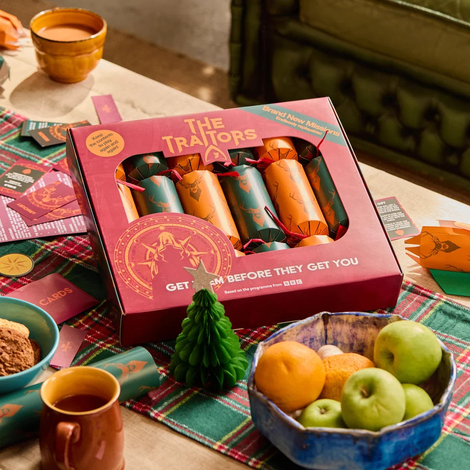 A boxed Christmas-themed card and cracker gift set labeled 'The Traitors' sits on a table surrounded by holiday props, including fruit, mugs, and a plaid runner. The box contains wrapped crackers and cards, with visible orange and teal wrapping and red accents.