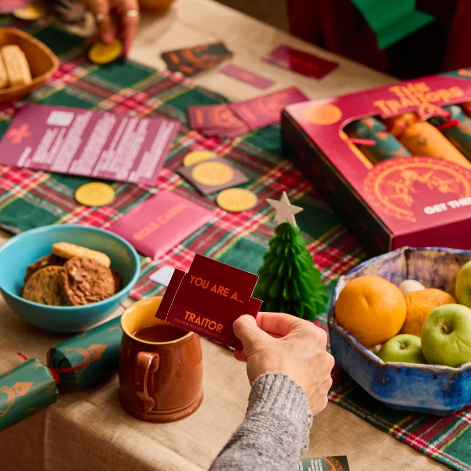 A person holds a red card in a holiday-themed game setup with a plaid tablecloth, game pieces, cookies, fruit, and a box labeled 'The Truth or Lies'.