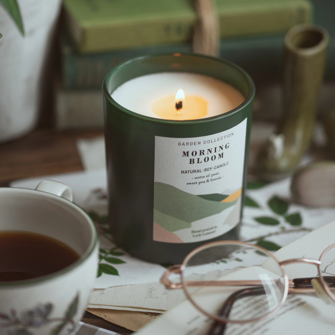 A dark green candle jar labeled 'GARDEN COLLECTION MORNING BLOOM' with a lit flame, styled on a tabletop with a cup of tea, glasses, and books in the background.