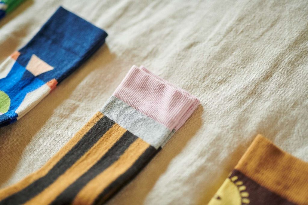A close-up of a folded knit sock on a textured cream fabric background. The sock features horizontal stripes in mustard yellow, black, and light gray, with a ribbed light pink cuff.