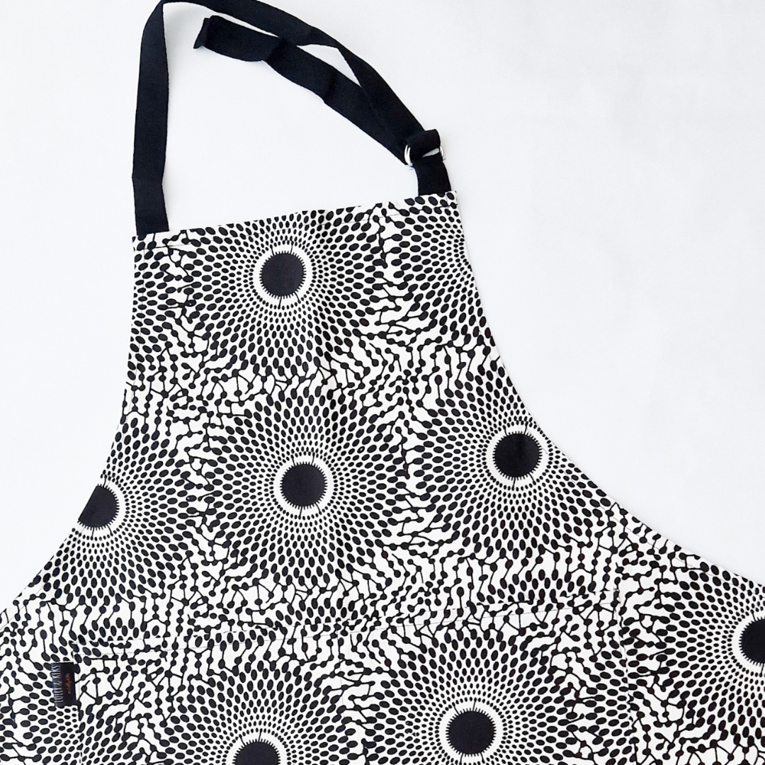 Black and white apron with a repeating sunburst pattern.  Features black straps and a small pocket.