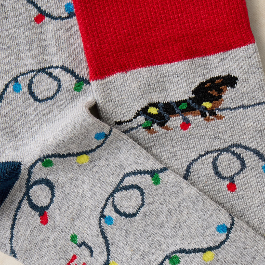 Close-up of a pair of gray knit socks with a red cuff, featuring a dachshund wrapped in colorful Christmas lights and repeated car-and-light motifs.