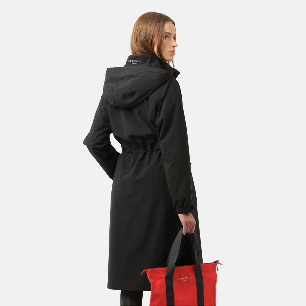 Black Ilse Jacobsen raincoat with hood, cinched waist, and long sleeves. Model holds a red Ilse Jacobsen tote bag.