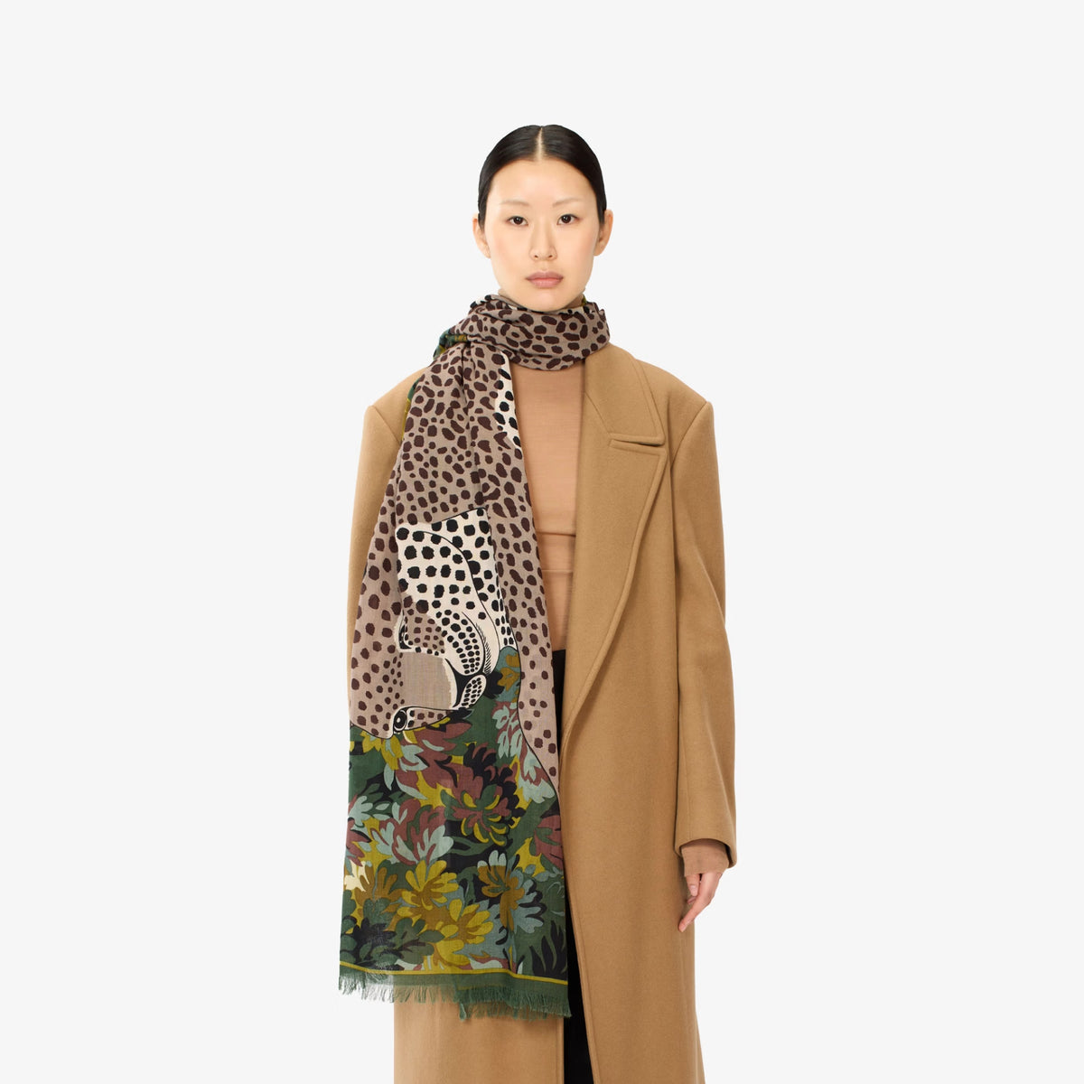 A model wearing a long camel coat, tan top, and a large leopard-print scarf with a floral section and elephant motif, photographed against a plain light gray background.