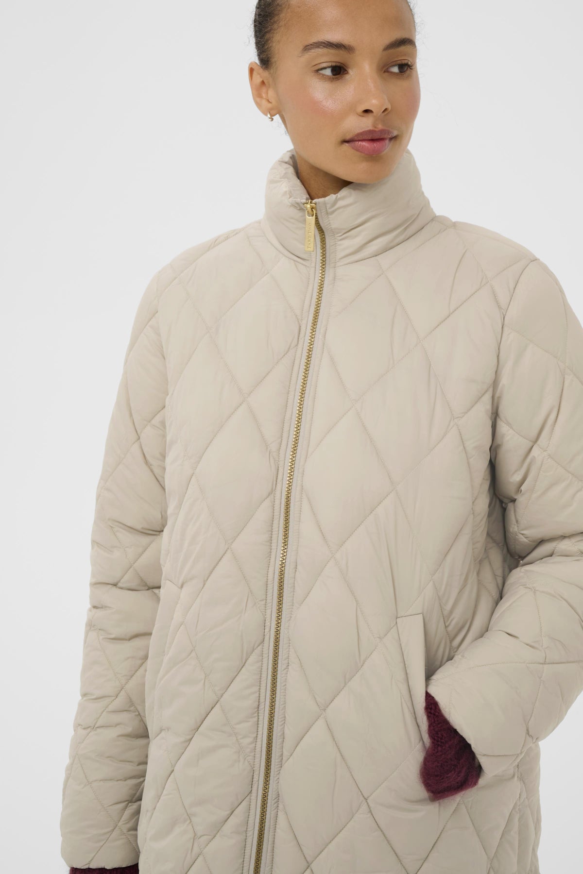 A woman wearing a quilted puffer jacket in a light beige/cream color with a high stand-up collar and a gold zipper, shown against a plain light gray studio background.