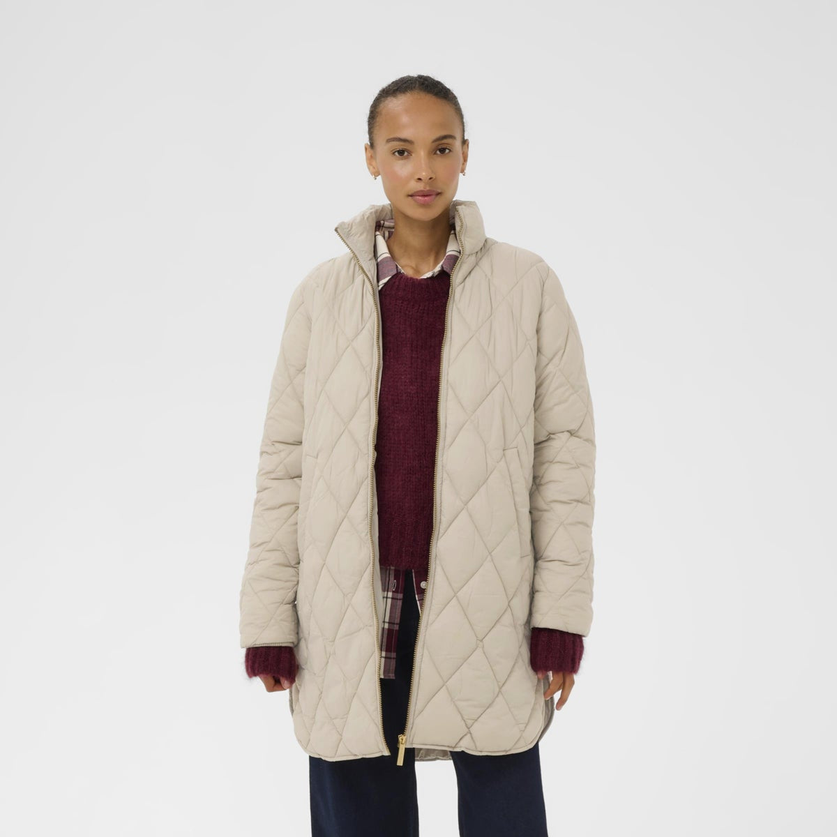 A model stands against a light gray studio background wearing a long quilted puffer coat in a light beige/cream color, layered over a burgundy sweater, plaid shirt, navy wide-leg pants, and black shoes.
