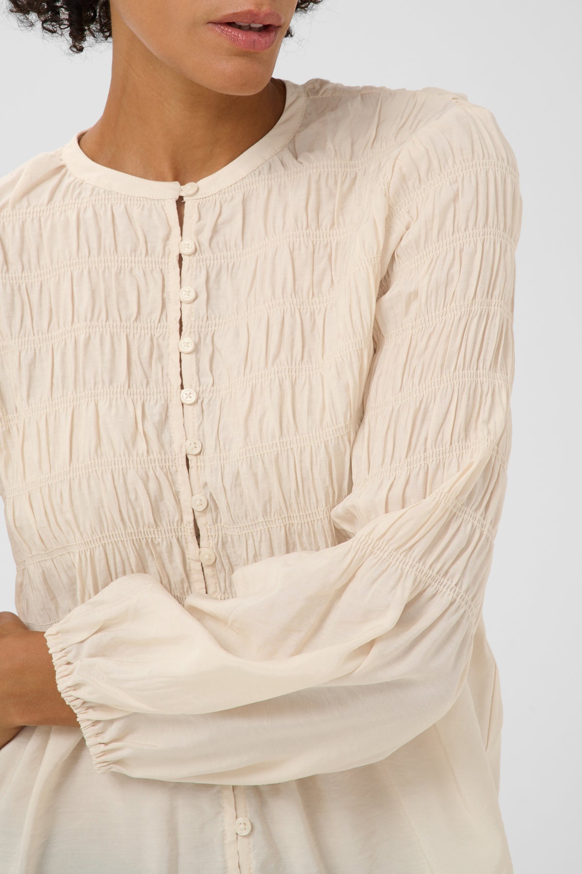 A close-up of a woman wearing a cream-colored, long-sleeved blouse with horizontal smocking and a button placket.