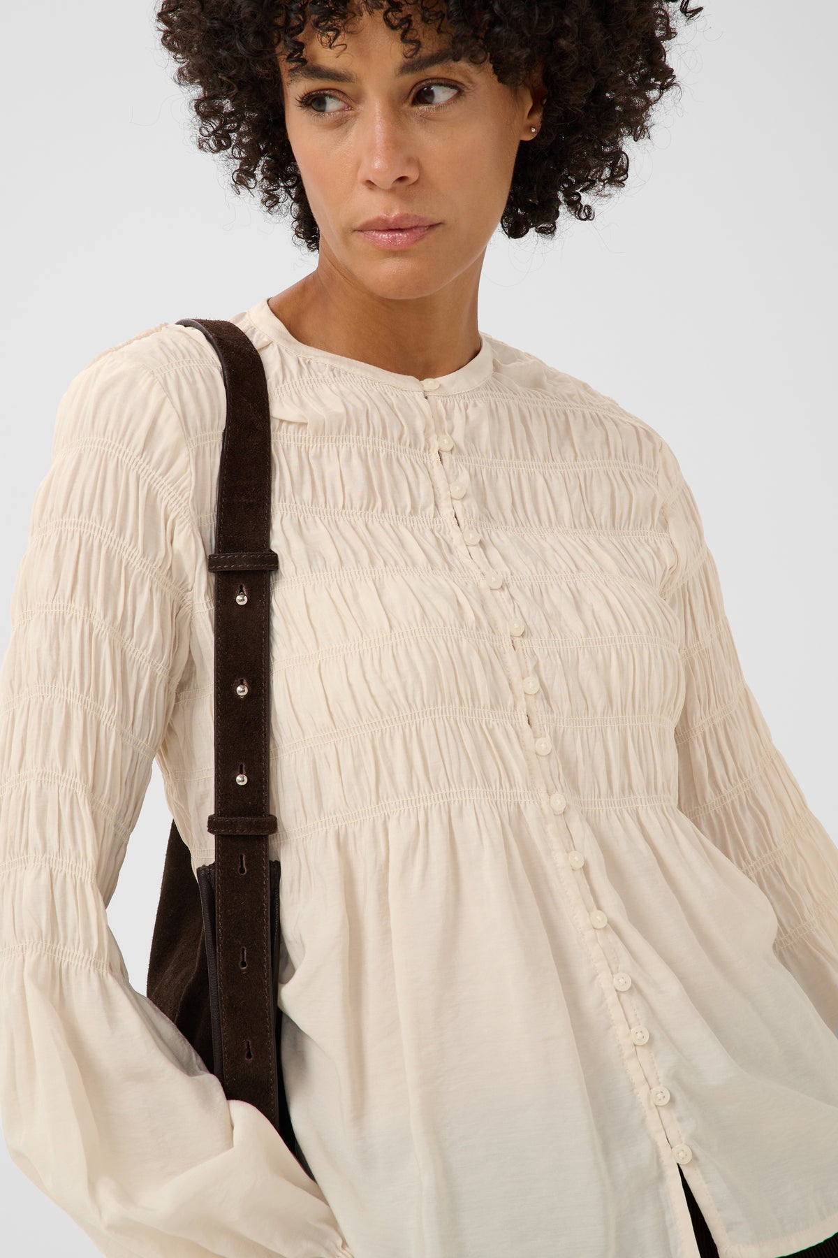 A woman wearing a cream-colored, long-sleeved smocked blouse with decorative buttons down the front, styled with a dark brown shoulder bag against a light gray background.