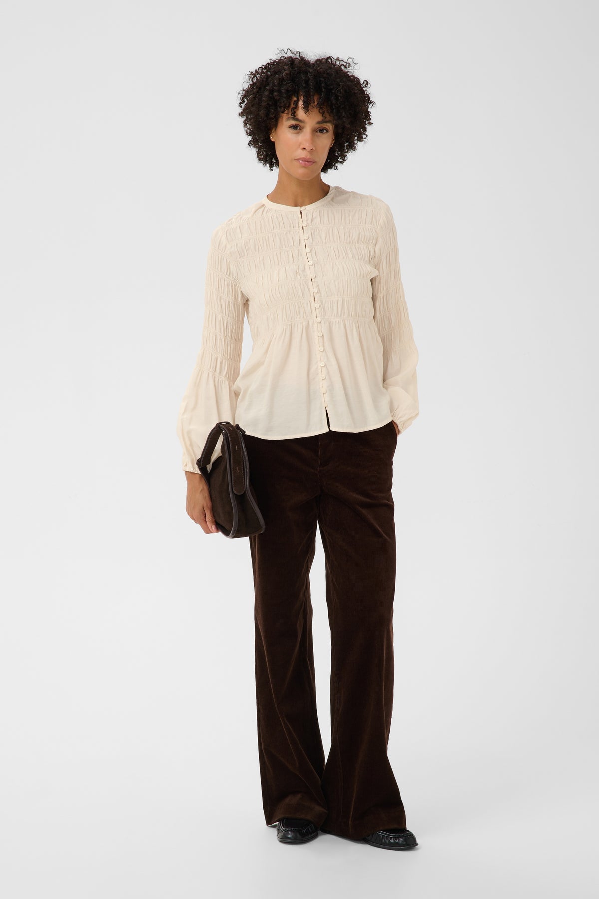 A model stands against a light gray studio background wearing a cream-colored, long-sleeved, button-front crinkled blouse and dark brown wide-leg trousers. She is holding a small black handbag.