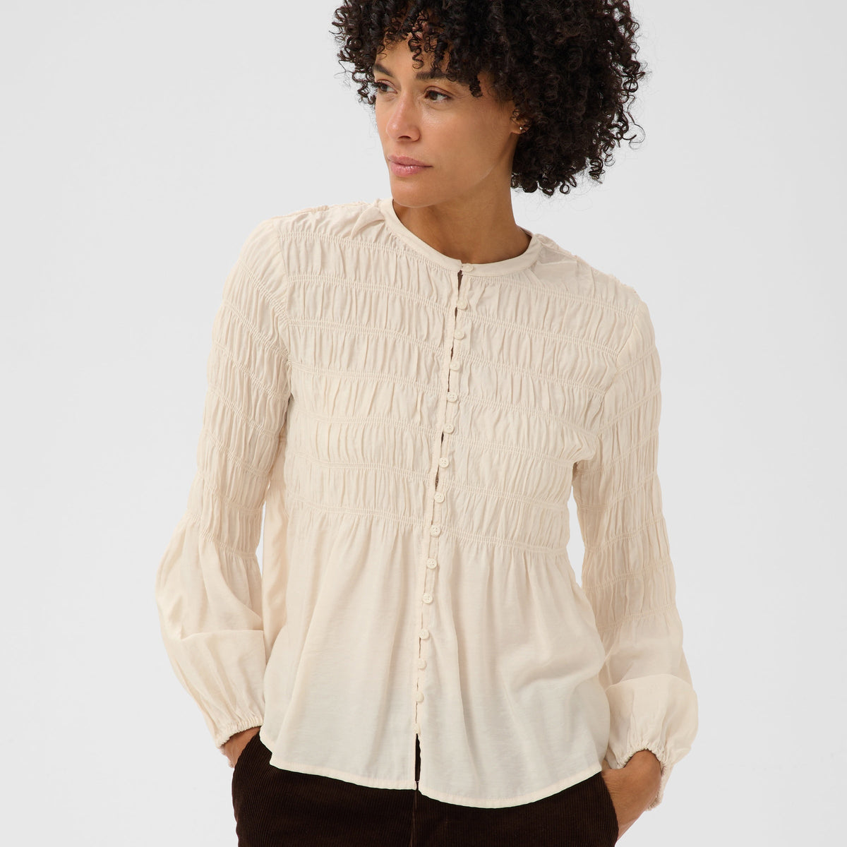 A woman with curly black hair wearing a cream-colored, long-sleeved, smocked blouse and dark brown pants, standing against a light gray studio background.