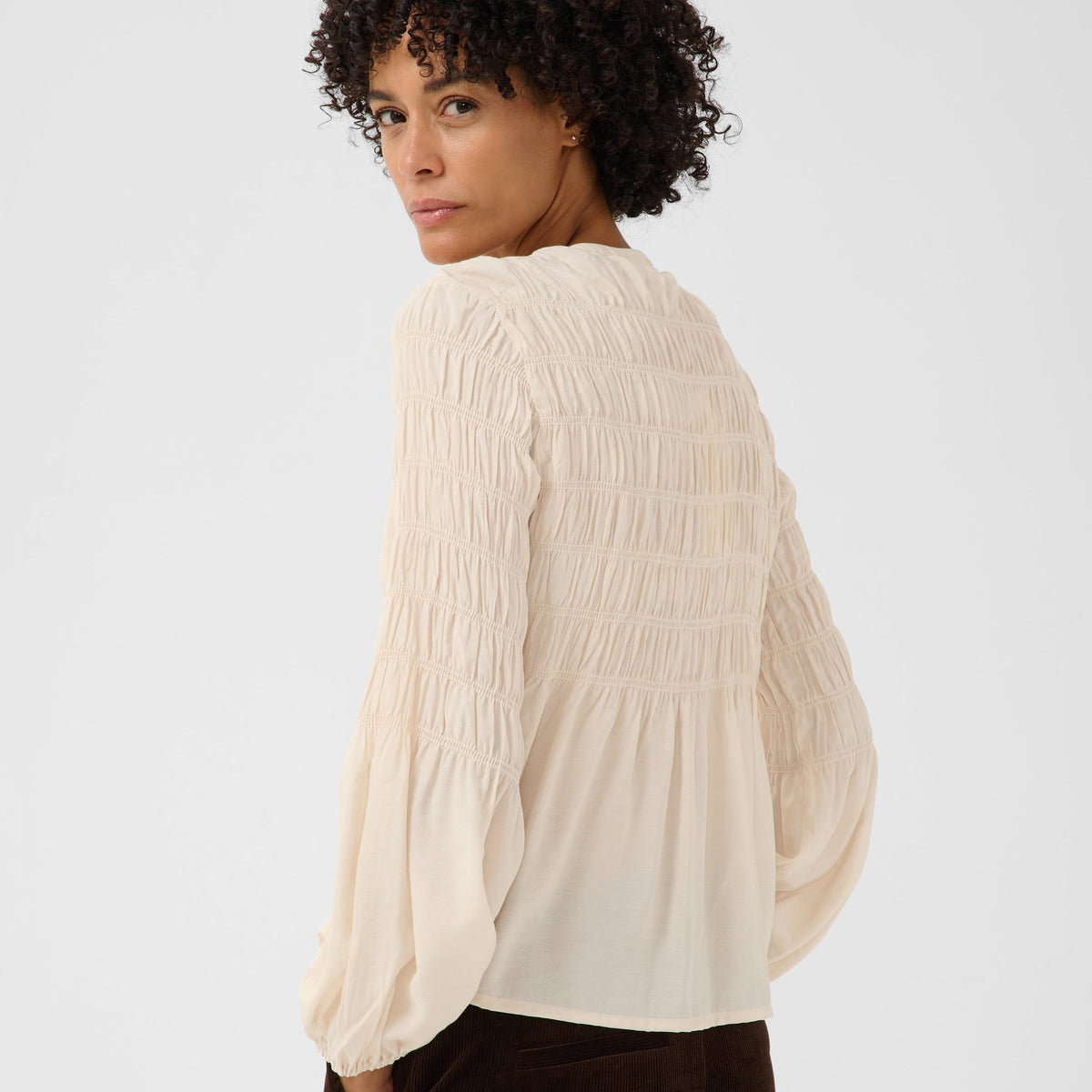 A woman with curly dark hair is shown from a side-back angle wearing a cream-colored, long-sleeved smocked blouse and dark brown corduroy pants against a light gray studio background.