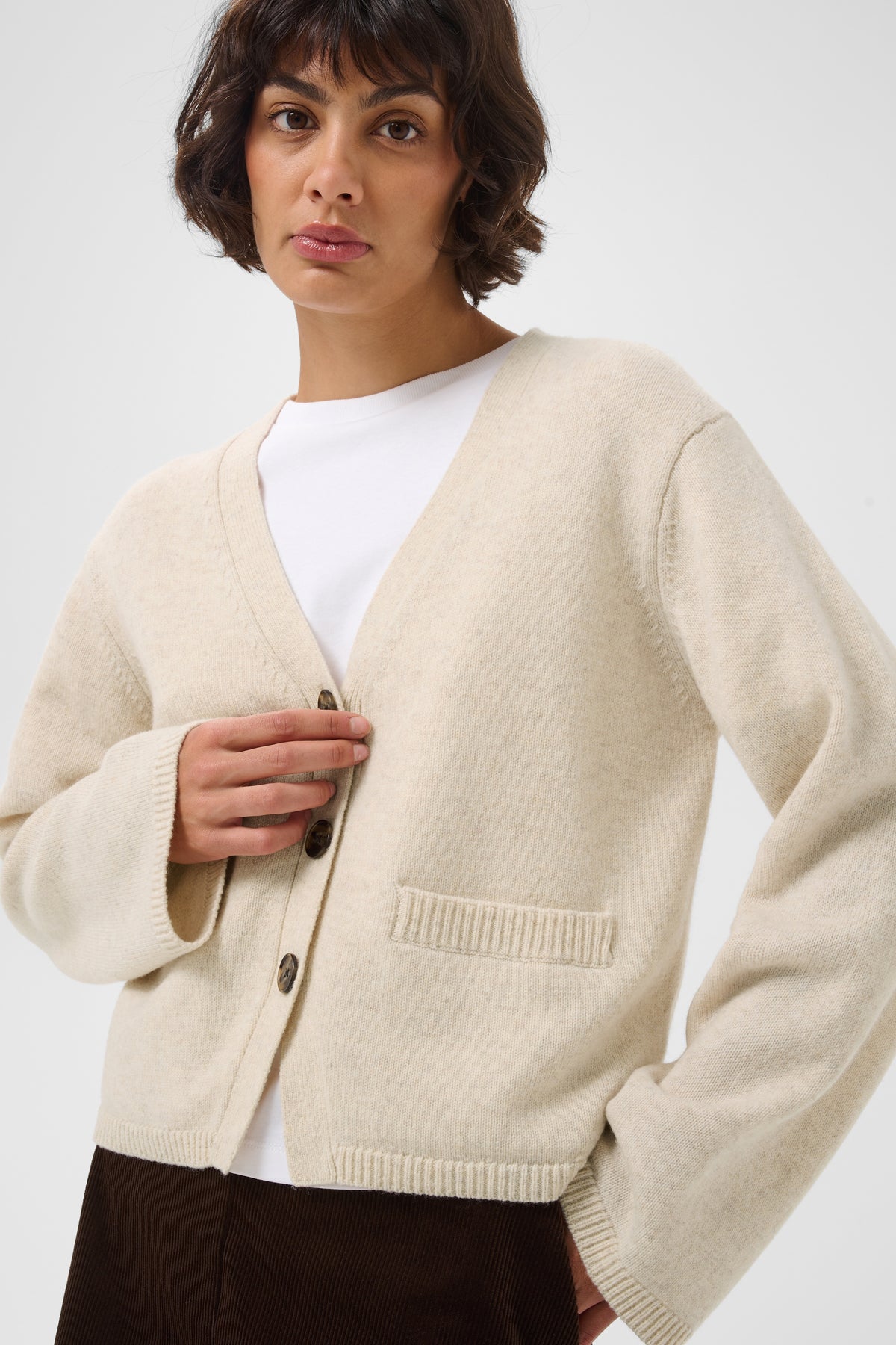 A woman wearing a cream-colored knit cardigan over a white top and dark brown trousers, photographed against a plain light gray studio background.