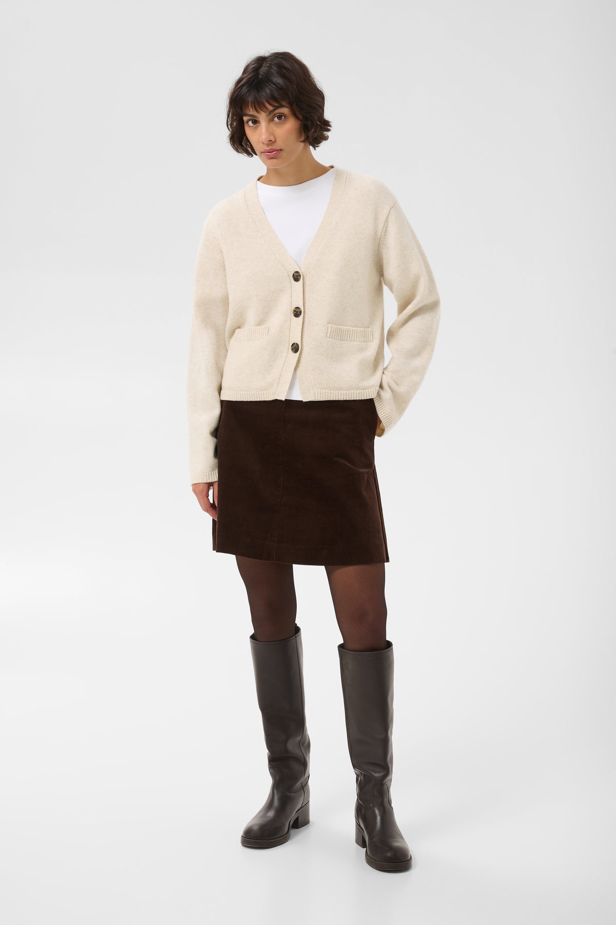 A model stands against a light gray studio background wearing a cream cardigan over a white top, a dark brown corduroy mini skirt, sheer dark tights, and dark brown knee-high boots.