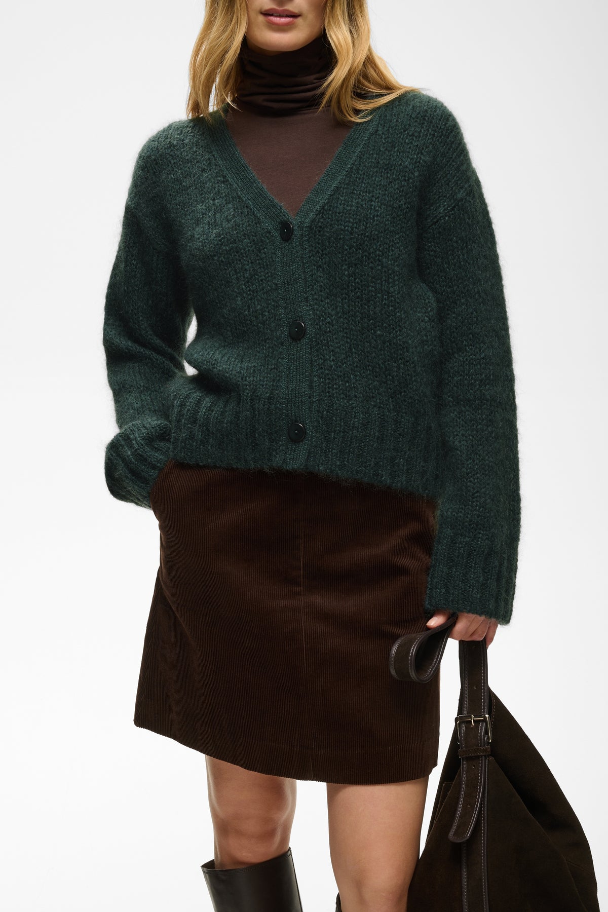 A woman wearing a dark green fuzzy knit cardigan over a dark brown turtleneck and a dark brown skirt, holding a dark brown tote bag against a plain white background.